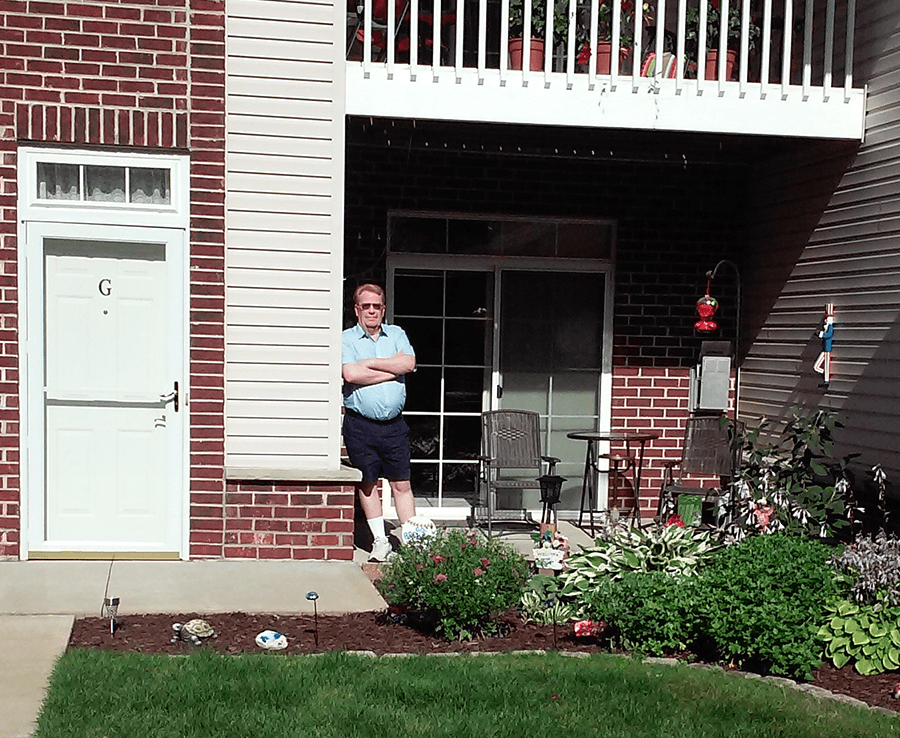 Henry stands on the porch of his condo. It's a red brick building with areas of white vinyl siding. His porch is behind low shrugs and is decorated with a wrought-iron bistro set and a hummingbird feeder.