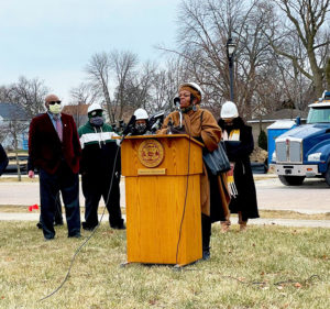 Great Midwest Bank Loan Officer Brenda Brown speaks outside behind a wooden podium, wearing a winter coat and a hard hat.