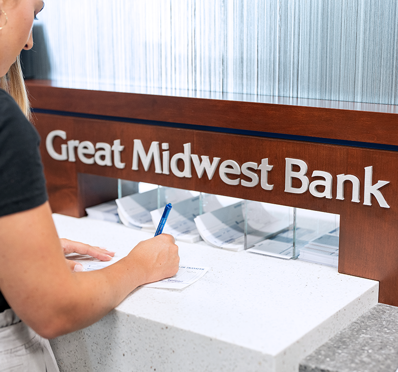 Young woman filling out a deposit slip on a tall table with lettering that reads "Great Midwest Bank."