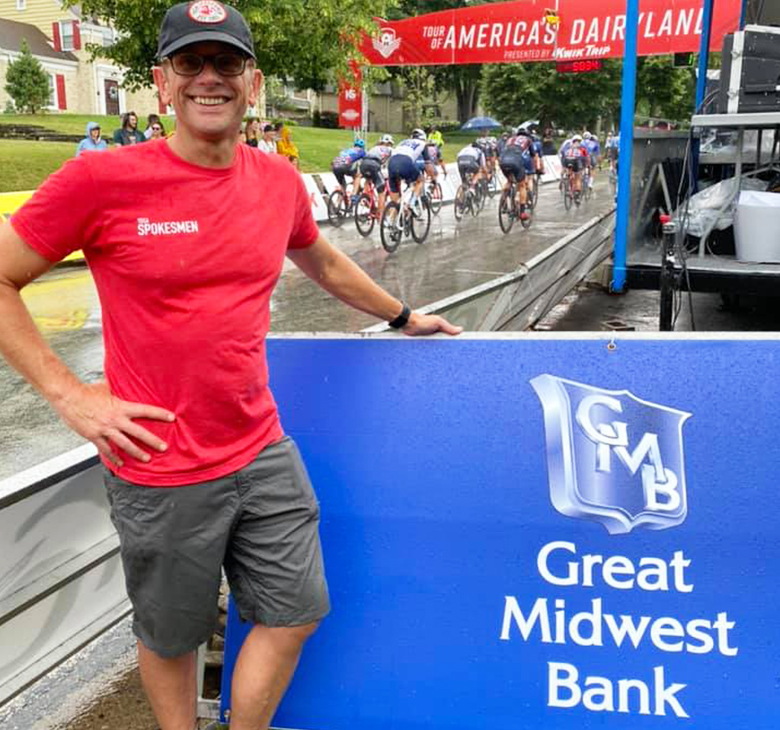 Great Midwest Bank Senior Vice President of Mortgage Operations stands in shorts and a tshirt next to a sign with the Great Midwest Bank logo on it. Behind him cyclists compete in a Tour of American's Dairyland race.