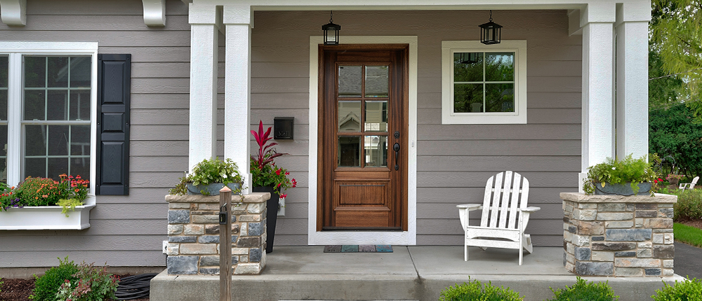 A white adirondack chair sits on the concrete front porch of a grey sided house with a wooden front door. The porch is flanked by white columns with stone bases.