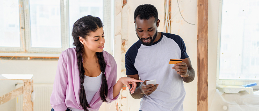 A young man and young woman stand in a room that has exposed beams, drywall, and wires. The man is holding a credit card, and both are looking at his smart phone.