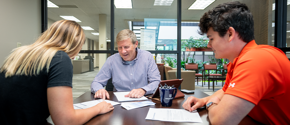 Great Midwest Bank loan officer John Hugo sits across from two first-time homebuyers while reviewing paperwork at a table.