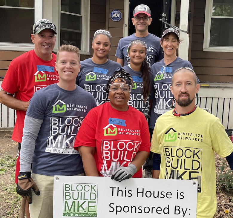 A group of Great Midwest Bank employees stand in Block Build MKE tshirts and safety gear around a sign that reads "This House is Sponsored by Great Midwest Bank"