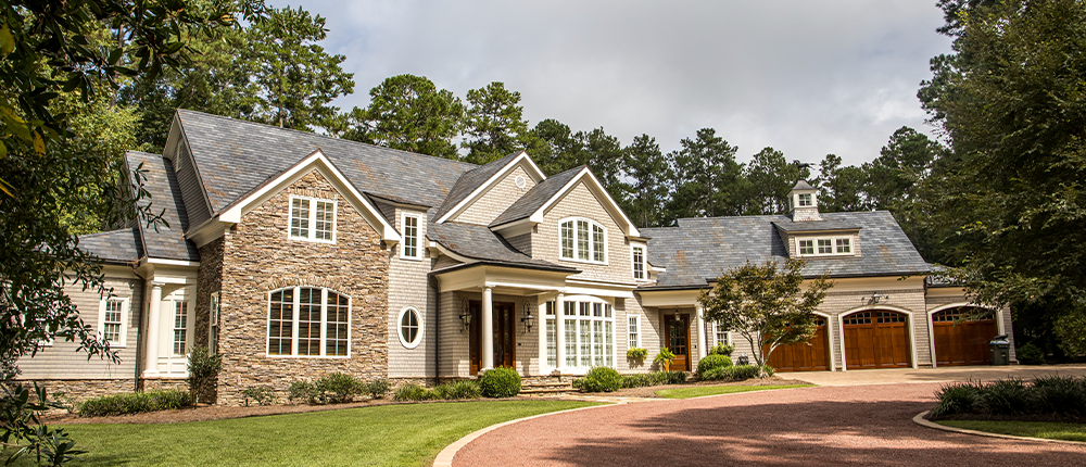 Large multi-story home with tan siding and stonework facade, clean landscaping, and a three-car attached garage sits in front of a circular red-bricked driveway.