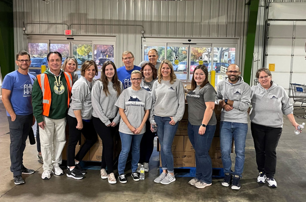 A group of 13 Great Midwest bank employees stand together in a warehouse at Feeding America Eastern Wisconsin.